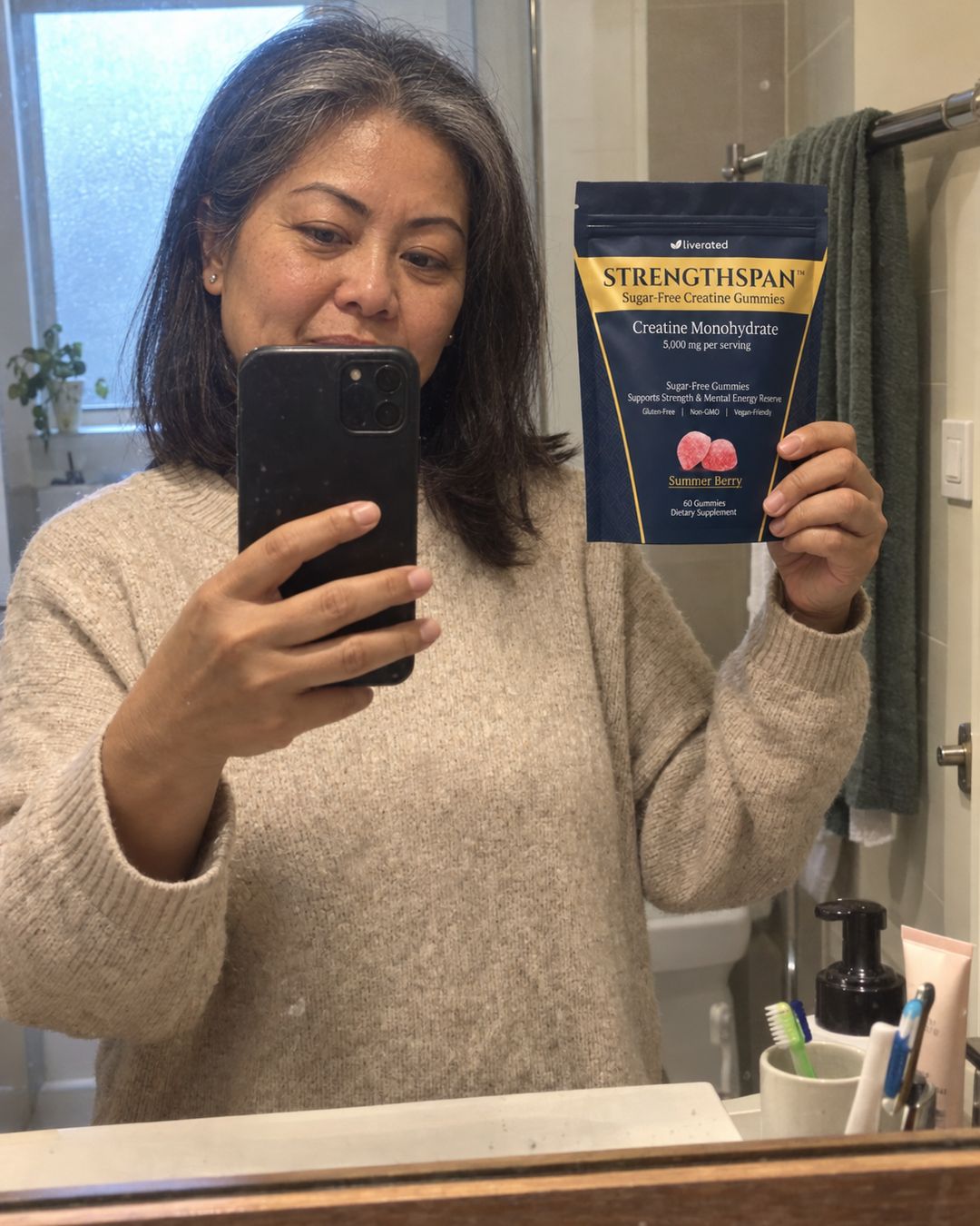 Photograph: a woman taking a mirror selfie in a hallway bathroom, holding a StrengthSpan pouch up beside her shoulder
