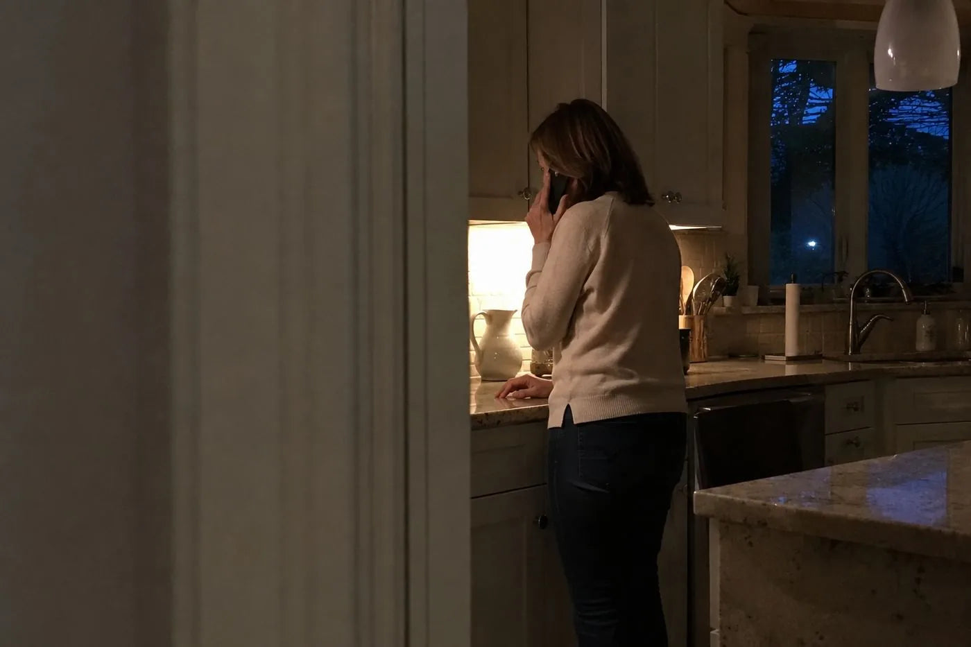 Seen through a half-open kitchen door: his wife on the phone at the counter at dusk