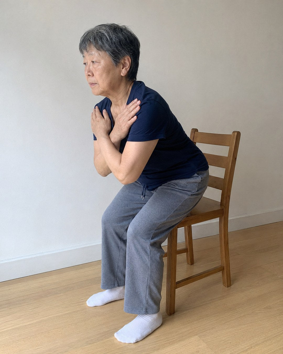 Photograph: a woman around 60 mid-stand from a plain wooden chair, arms crossed over her chest, feet flat